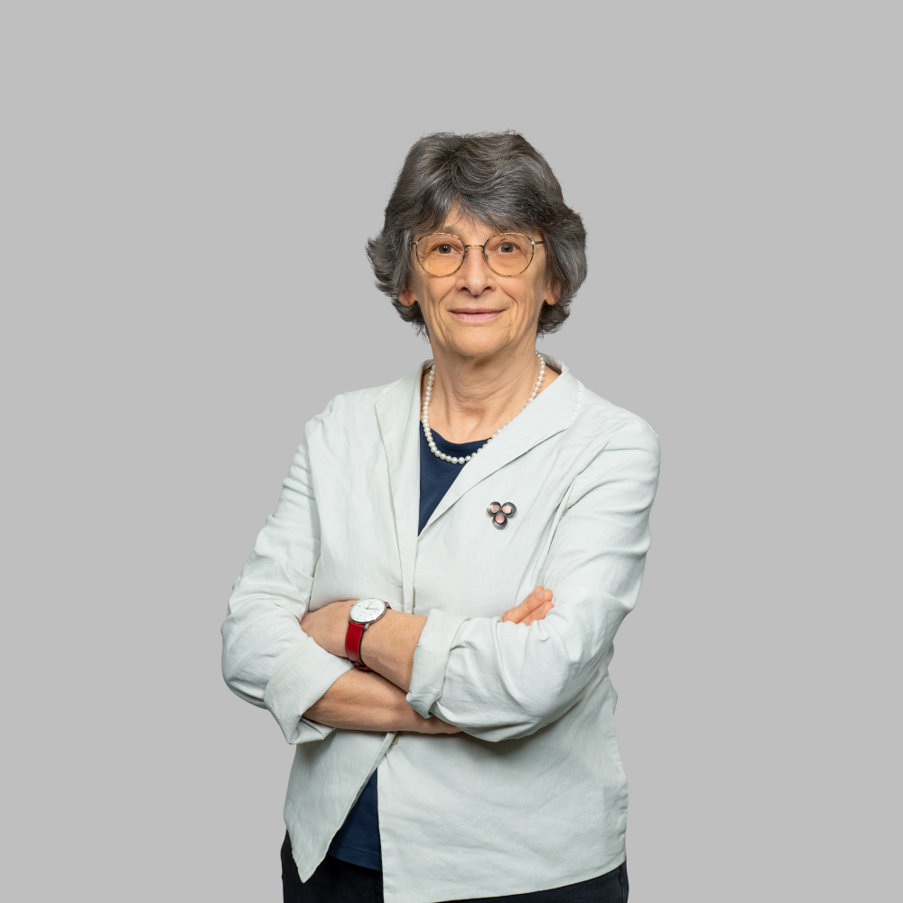 Portrait von Prof. Dr. Elvira Glaser, weiblich lesbare Person mit kurzen, dunkelgrauen Haaren, Brille, Perlenkette, blauem Oberteil und mintfarbenem Blazer, daran eine Brosche, mit einer Armbanduhr am linken Arm