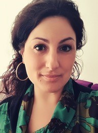 Portrait von Prof. Dr. Meldem Kulaçatan, weiblich lesbare Person mit dunklen, lockigen, langen Haaren, die zu einem Zopf gebunden sind, Ohrringen und bunt-gemusterter Bluse