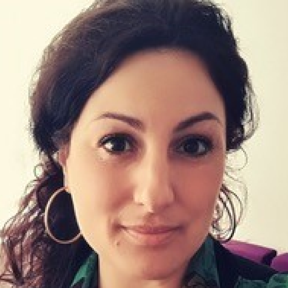 Portrait von Prof. Dr. Meldem Kulaçatan, weiblich lesbare Person mit dunklen, lockigen, langen Haaren, die zu einem Zopf gebunden sind, Ohrringen und bunt-gemusterter Bluse