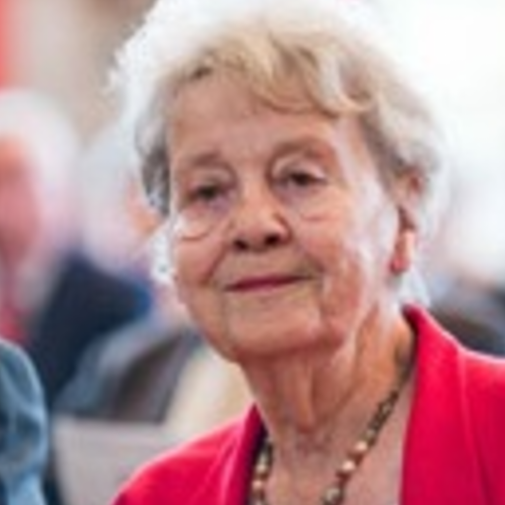 Portrait Inge Strauch, weiblich lesbare, weisse Person mit kurzen, lockigen Haaren, rotem Oberteil und Kette