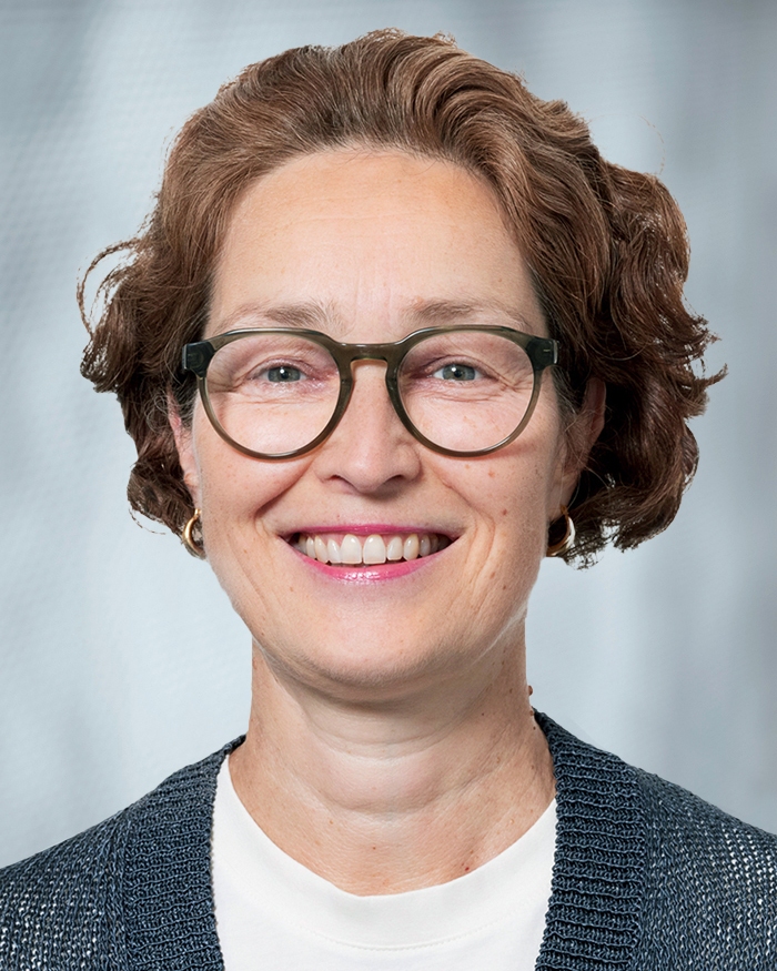 Portrait Tanja Neve-Seyfarth, weiblich lesbare, weisse Person mit mittellangem, lockigem Haar, Brille, Ohrringen, weissem Oberteil und grauer Jacke